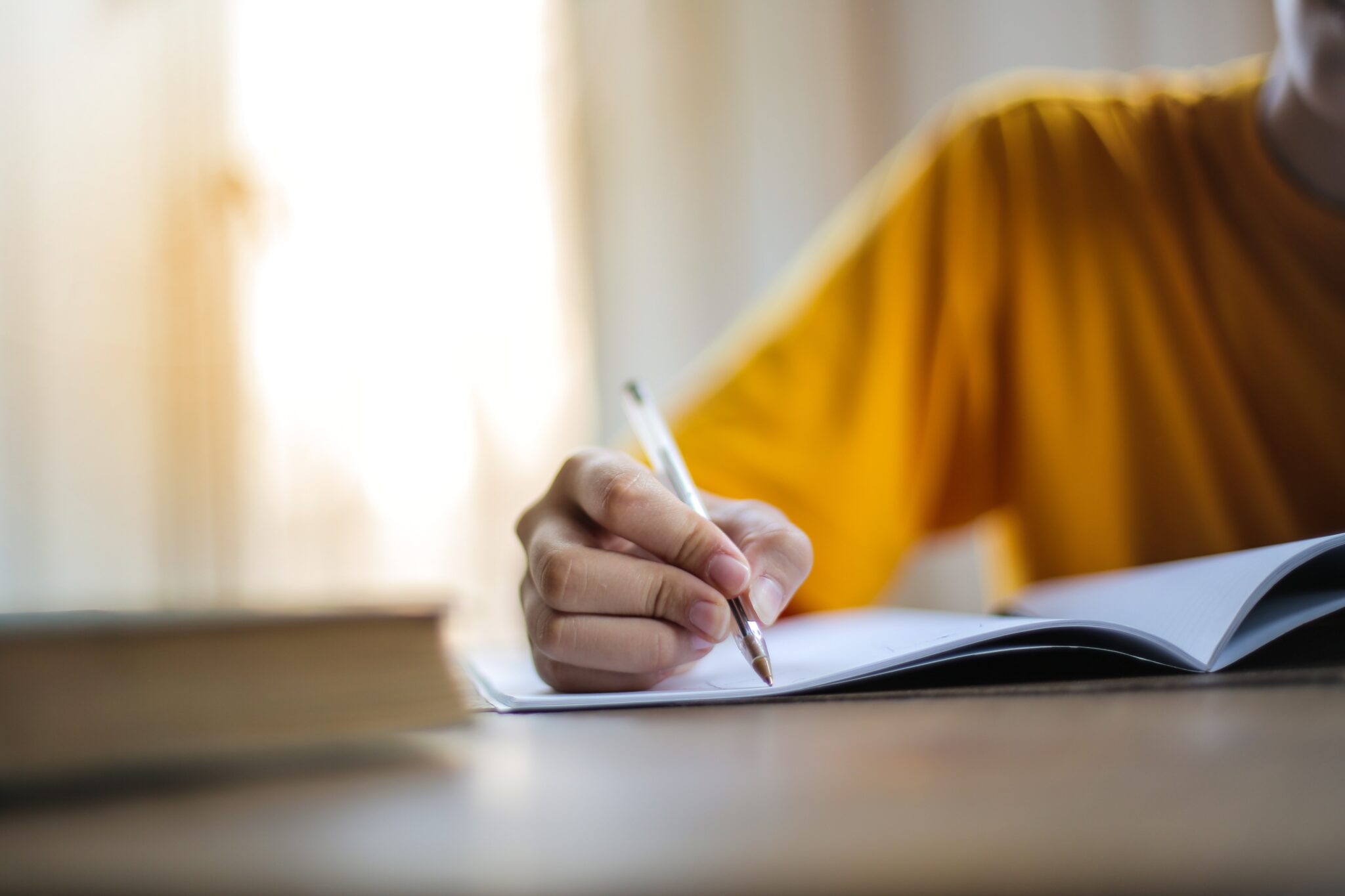 Transcreators are often copywriters experienced in marketing. Description: a person in a yellow shirt writing with a pen in a notebook, which is on a desk.