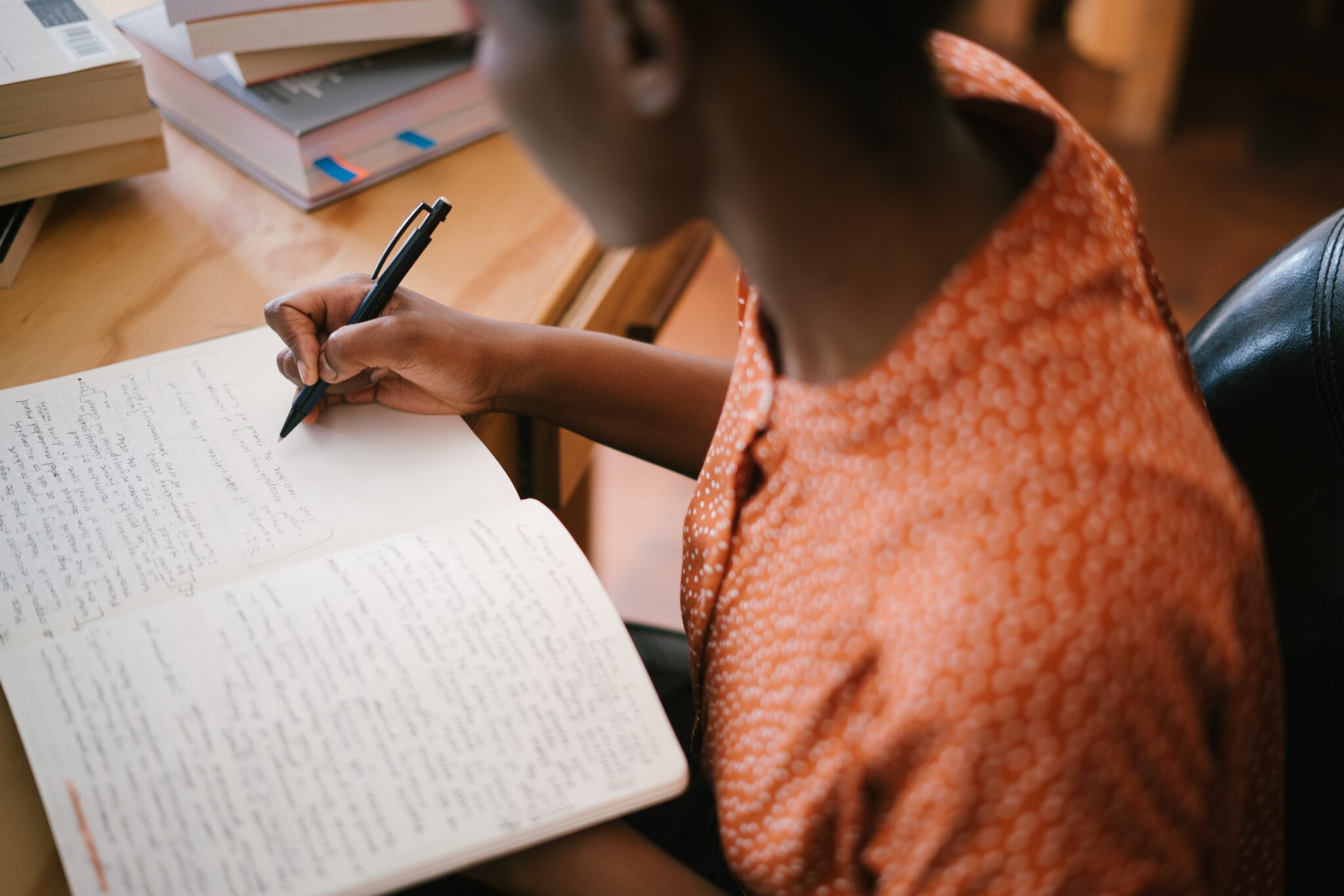 Transliteration takes characters from one alphabet and renders them into another.

Description: A woman wearing an orange blouse is writing in a notebook on her desk, surrounded by books.